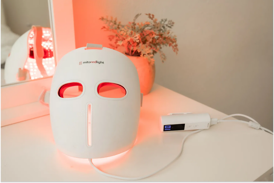 MitoRedLight red light therapy face mask glowing with red LED light, placed on a white surface with a controller device beside it.
