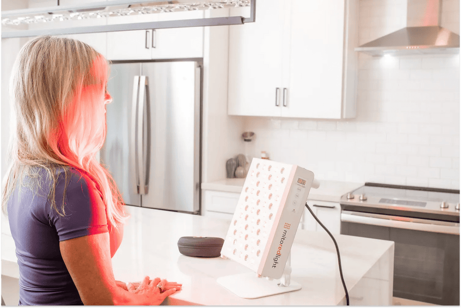 Woman sitting in front of a MitoRedLight red light therapy panel at home, using red light therapy for recovery.